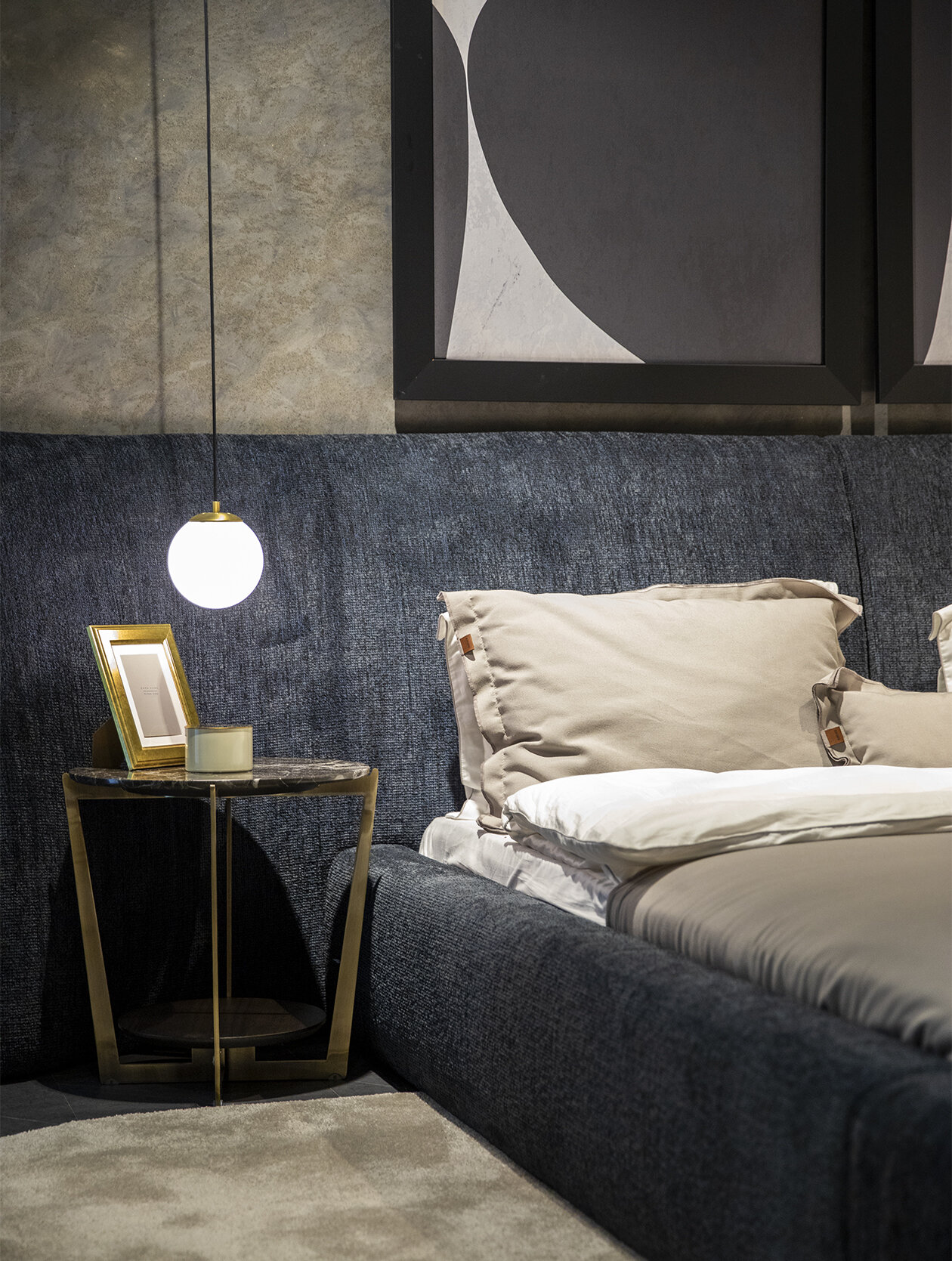Stylish bedroom with a dark upholstered headboard, modern abstract wall art, and a cozy nightstand featuring a gold frame, a candle, and a framed photo, all emphasizing contemporary furniture design.