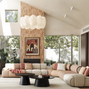 Elegant living room featuring a large, curved sectional sofa with beige and pink cushions, surrounded by natural light from expansive windows and decorated with modern art and a statement chandelier.