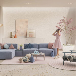 An elegant living room featuring a pastel blue sectional sofa, pink cushions, and a cozy pink area rug. A woman in a pink dress stands near a large window with sheer curtains, surrounded by stylish de.
