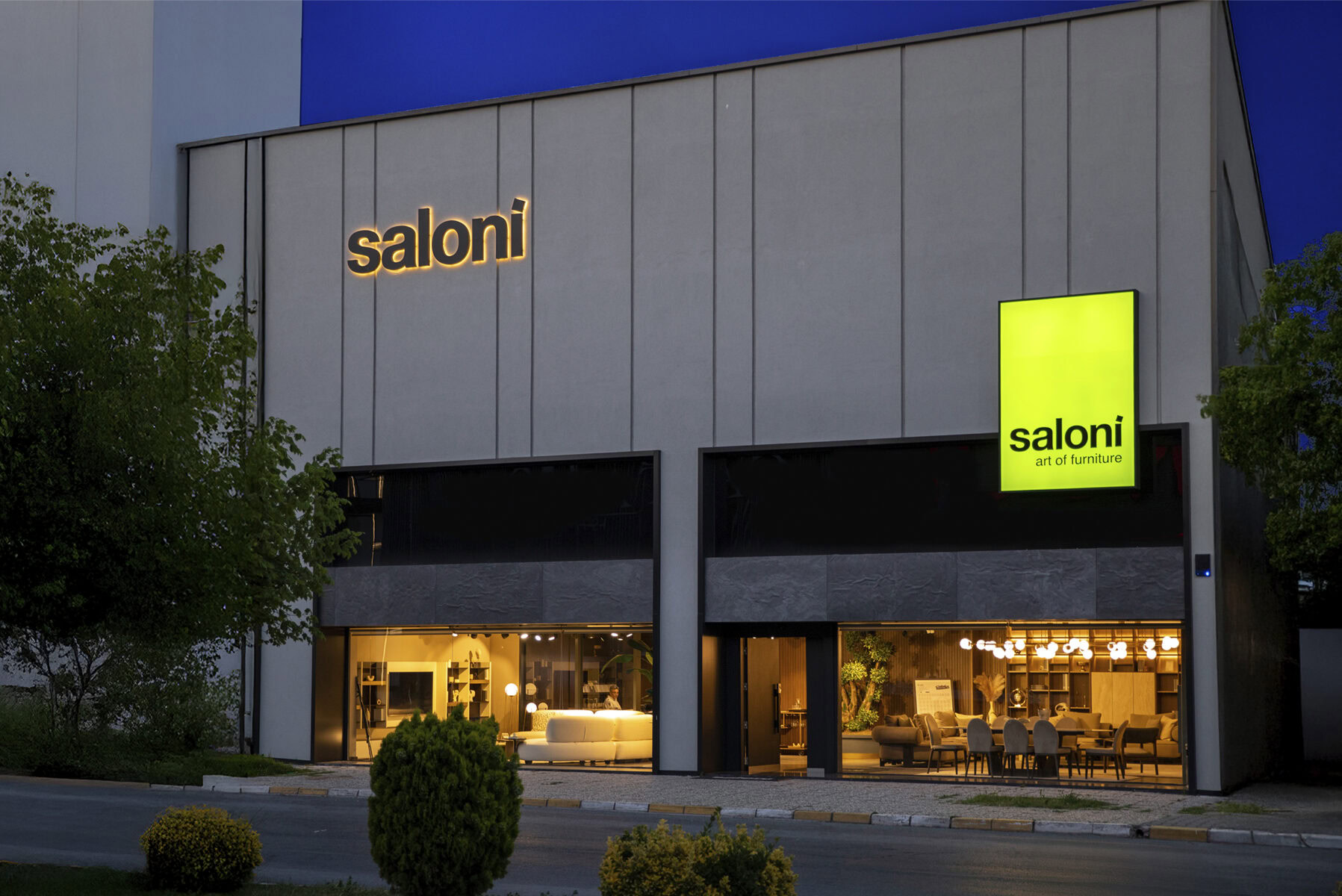 Modern furniture store exterior showcasing Saloni Furniture's logo illuminated at dusk, with a sleek facade and inviting interiors featuring stylish living room furniture displays.