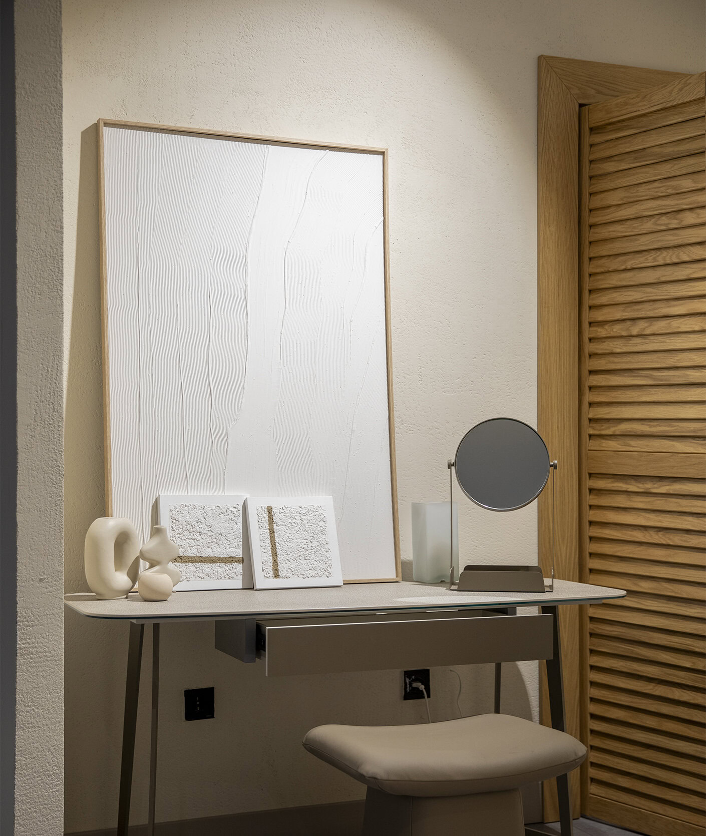 Sleek modern vanity desk with a round mirror, minimalist decor, and textured art pieces, perfect for stylish home office or bedroom setups. High-quality furniture from Saloni Furniture.